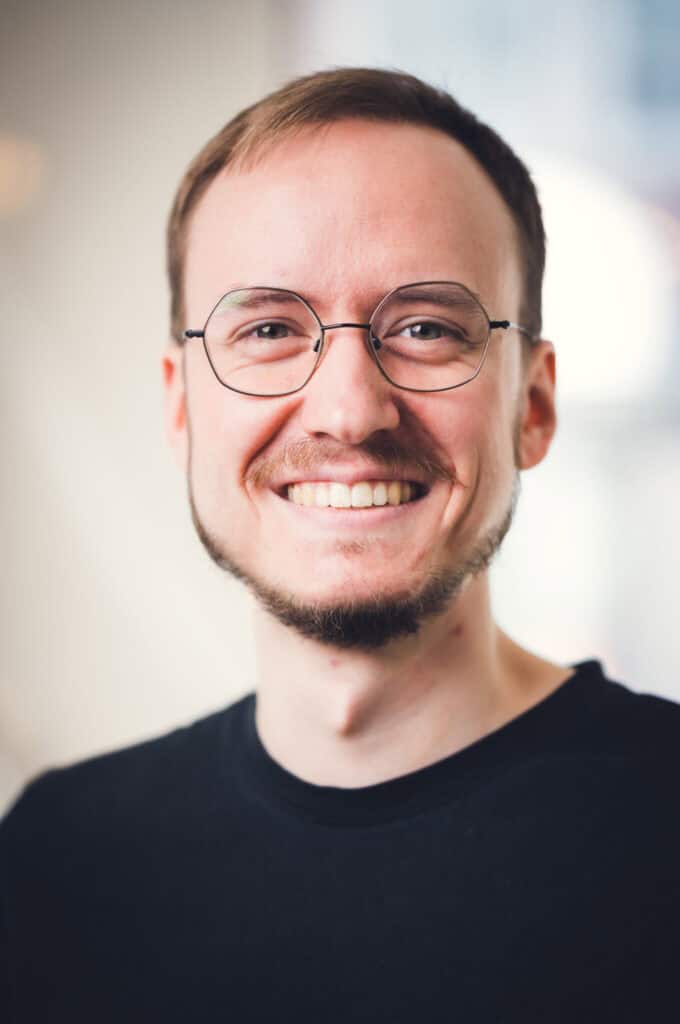 Junger Mann mit Brille und dunklem Haar, in schwarzem T-Shirt, vor hellem, unscharfem Hintergrund.