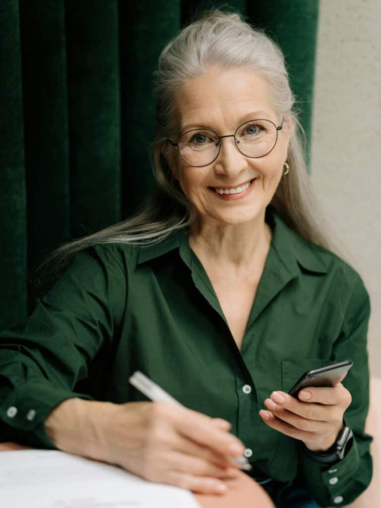 Eine stilvolle ältere Dame mit silbergrauem Haar und Brille sitzt lächelnd an einem Tisch. Sie trägt ein elegantes grünes Hemd und hält ein Smartphone in einer Hand, während sie mit der anderen handschriftliche Notizen macht. Der Hintergrund aus dunkelgrünen Vorhängen ergänzt das harmonische Farbschema.